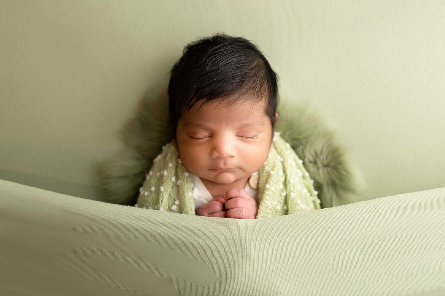 Newborn baby sleeping peacefully wrapped in a green knit blanket with fur trim against a soft green background.