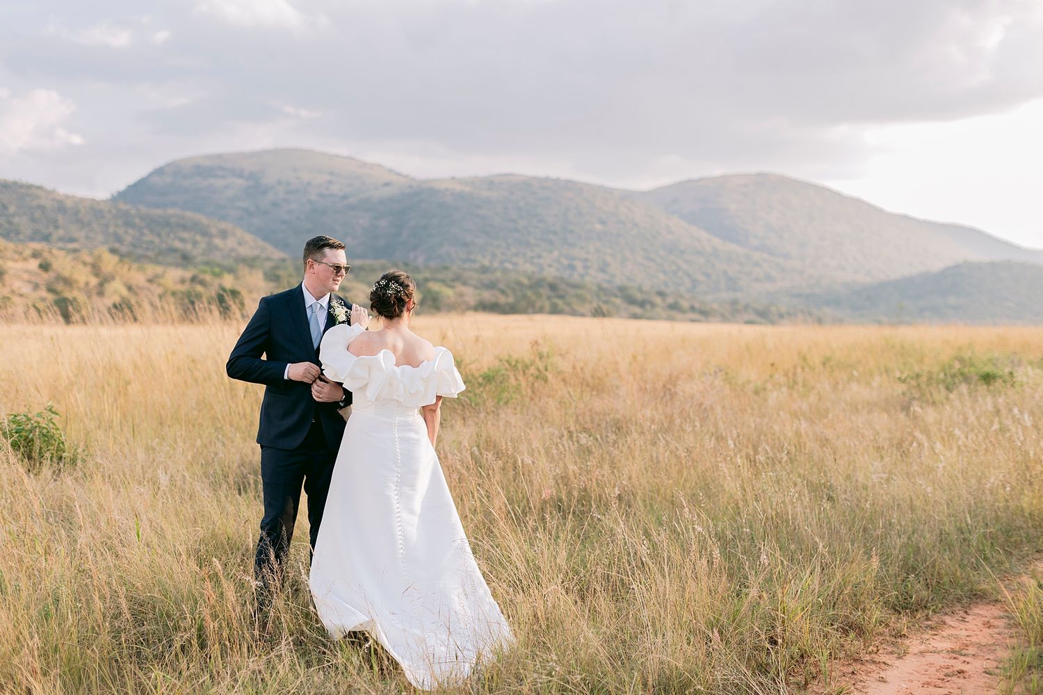 Bride and groom embrace in golden field with mountain backdrop on their wedding day.