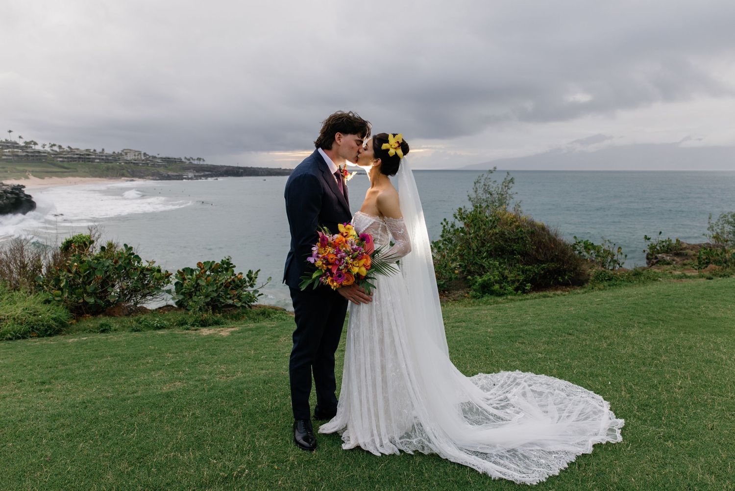 Bride and groom share a kiss on green lawn overlooking ocean beach on cloudy day, bride holds colorful bouquet.