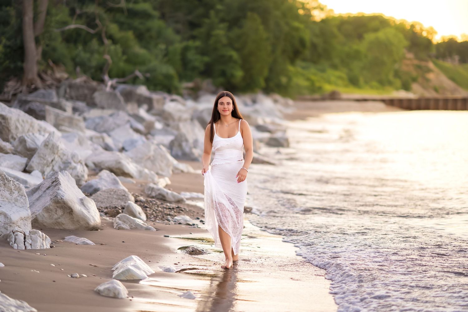 Woman in white dress walking along rocky beach shoreline at golden sunset.