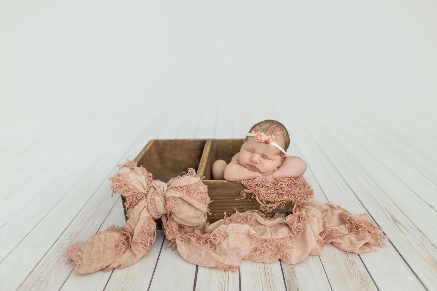 Newborn baby sleeping peacefully in a rustic wooden crate with soft pink gauze fabric on white wood floor.