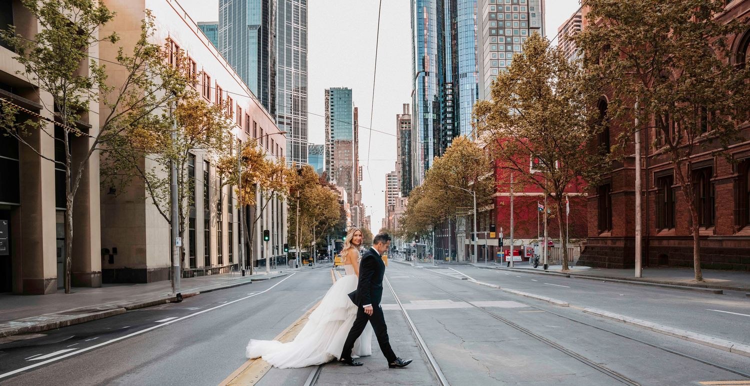 Bride and groom crossing an empty city street in wedding attire, with tall buildings and autumn trees lining the urban boulevard.