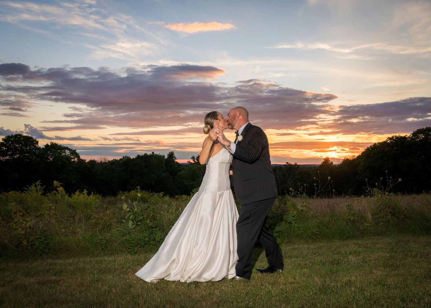 Bride and groom share a romantic kiss at sunset during outdoor wedding ceremony with colorful sky backdrop.