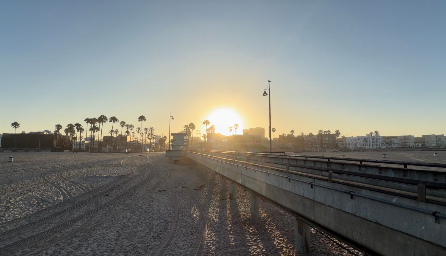 Sun setting over a beach pier with palm trees and golden sand in the background.