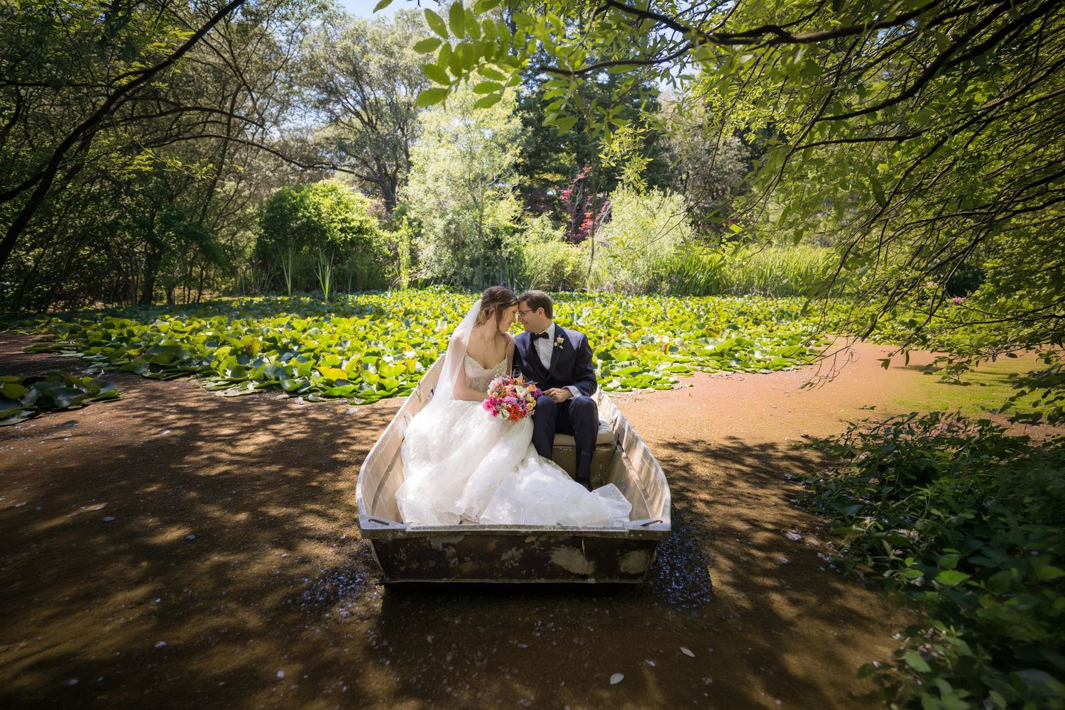 Bride and groom sitting in a decorative boat surrounded by lush greenery and lily pads in a romantic garden setting.