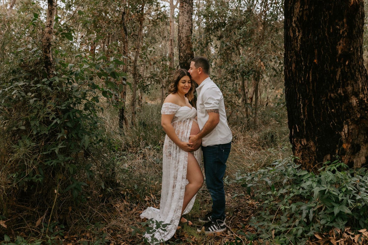 Couple posing in forest during maternity photoshoot, woman in white lace dress cradling baby bump surrounded by trees.