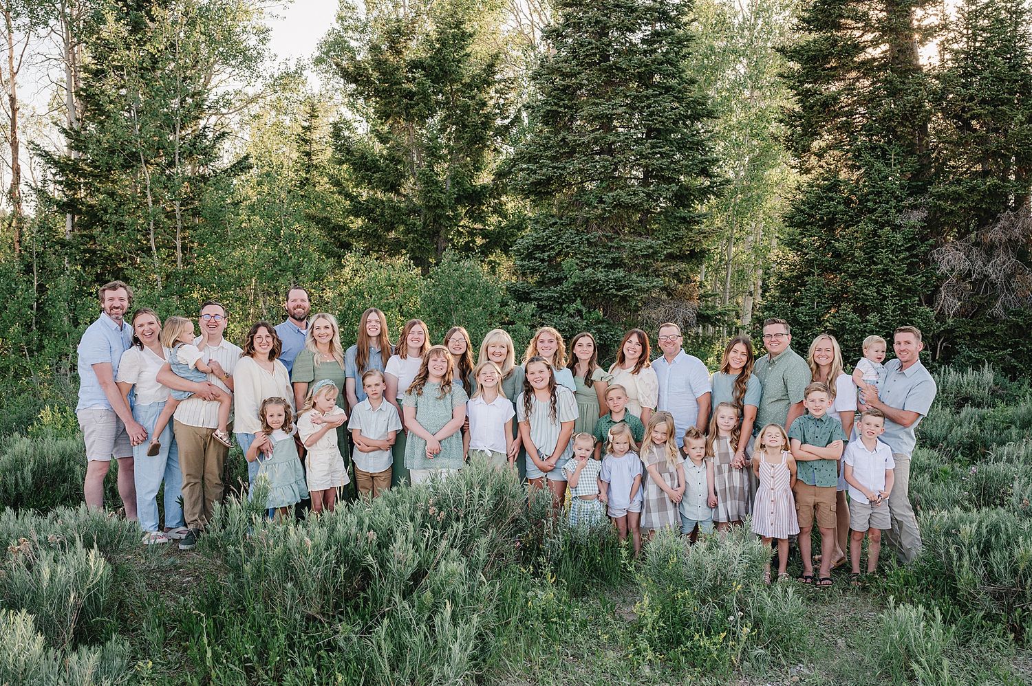 Large extended family group wearing coordinated neutral outfits poses together against an evergreen forest backdrop.
