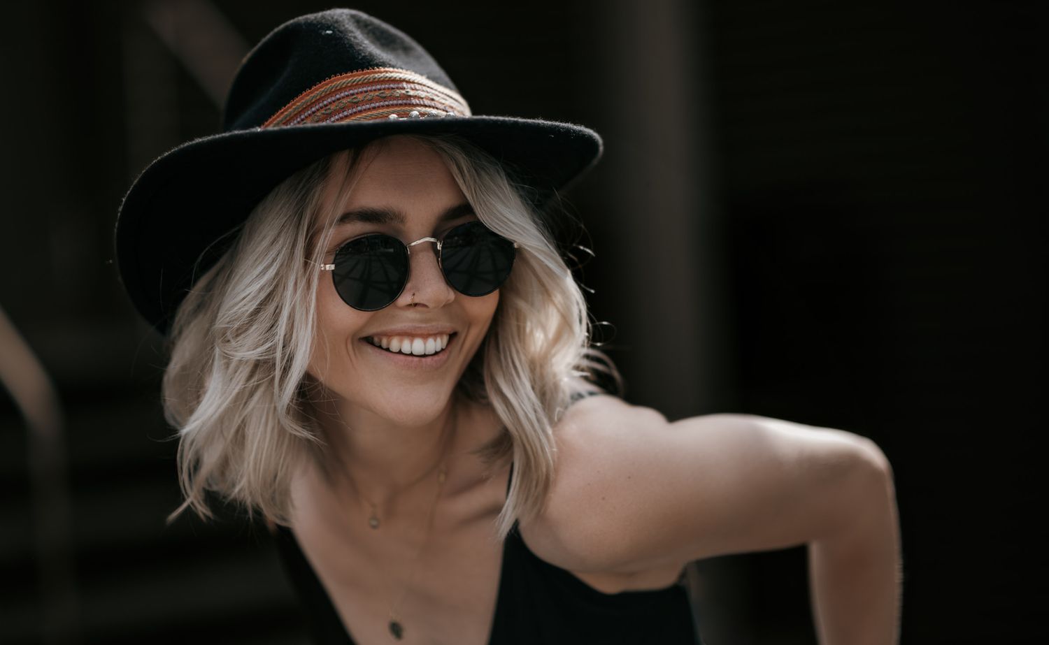 Smiling blonde woman wearing black hat and round sunglasses outdoors.