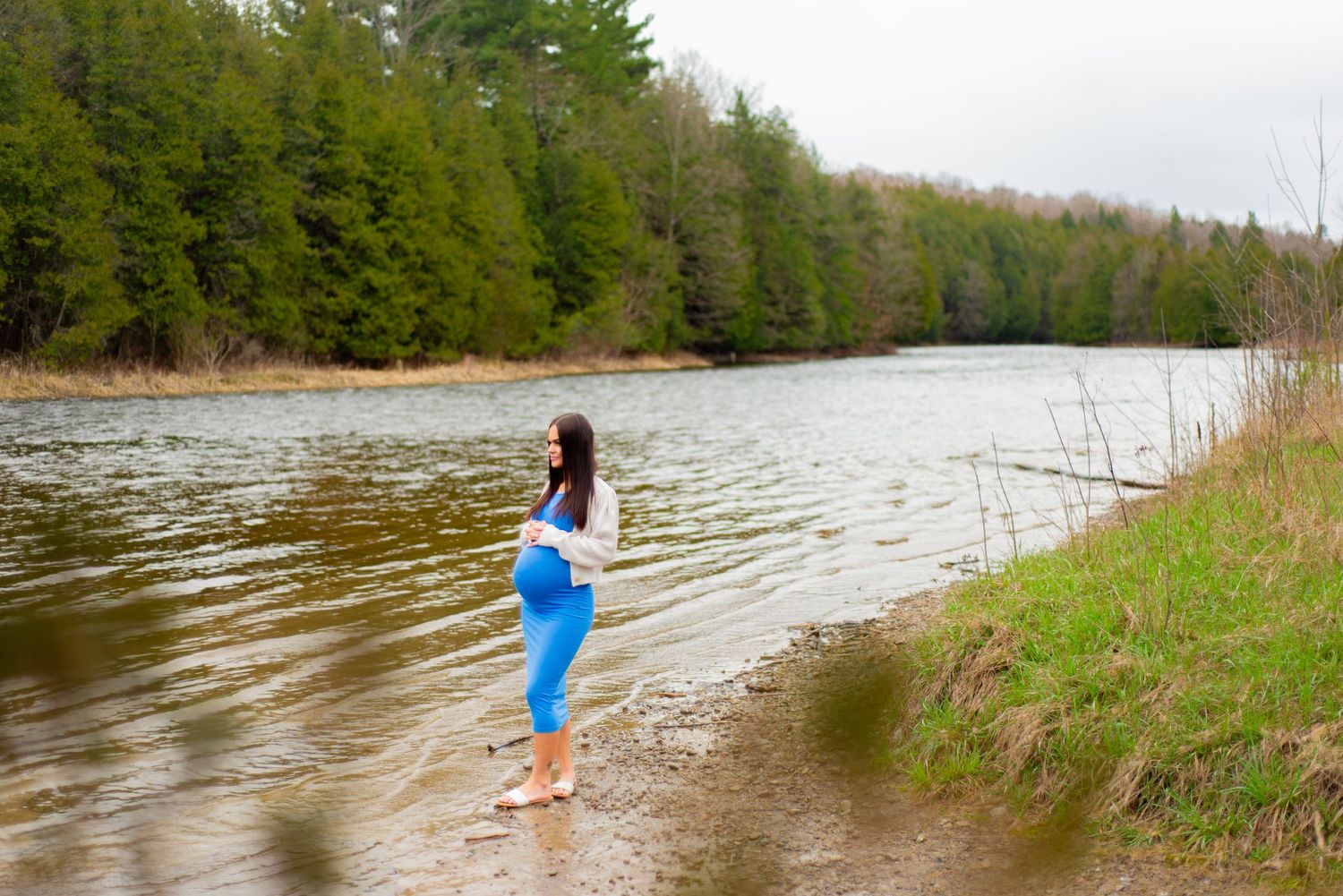 A person stands at the edge of a river surrounded by evergreen trees.