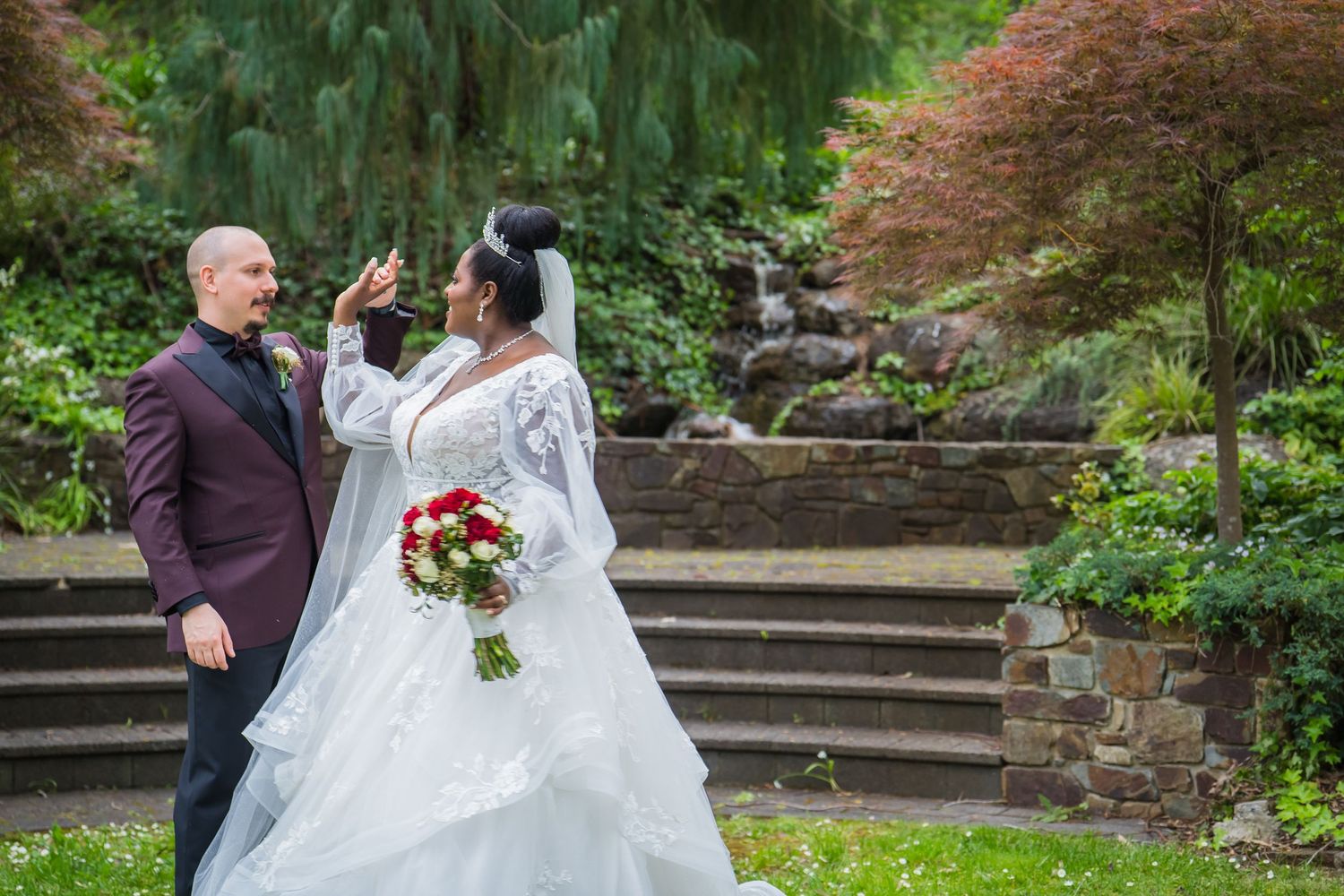 A couple shares a romantic moment in a lush garden setting with stone steps and vibrant greenery in the background.