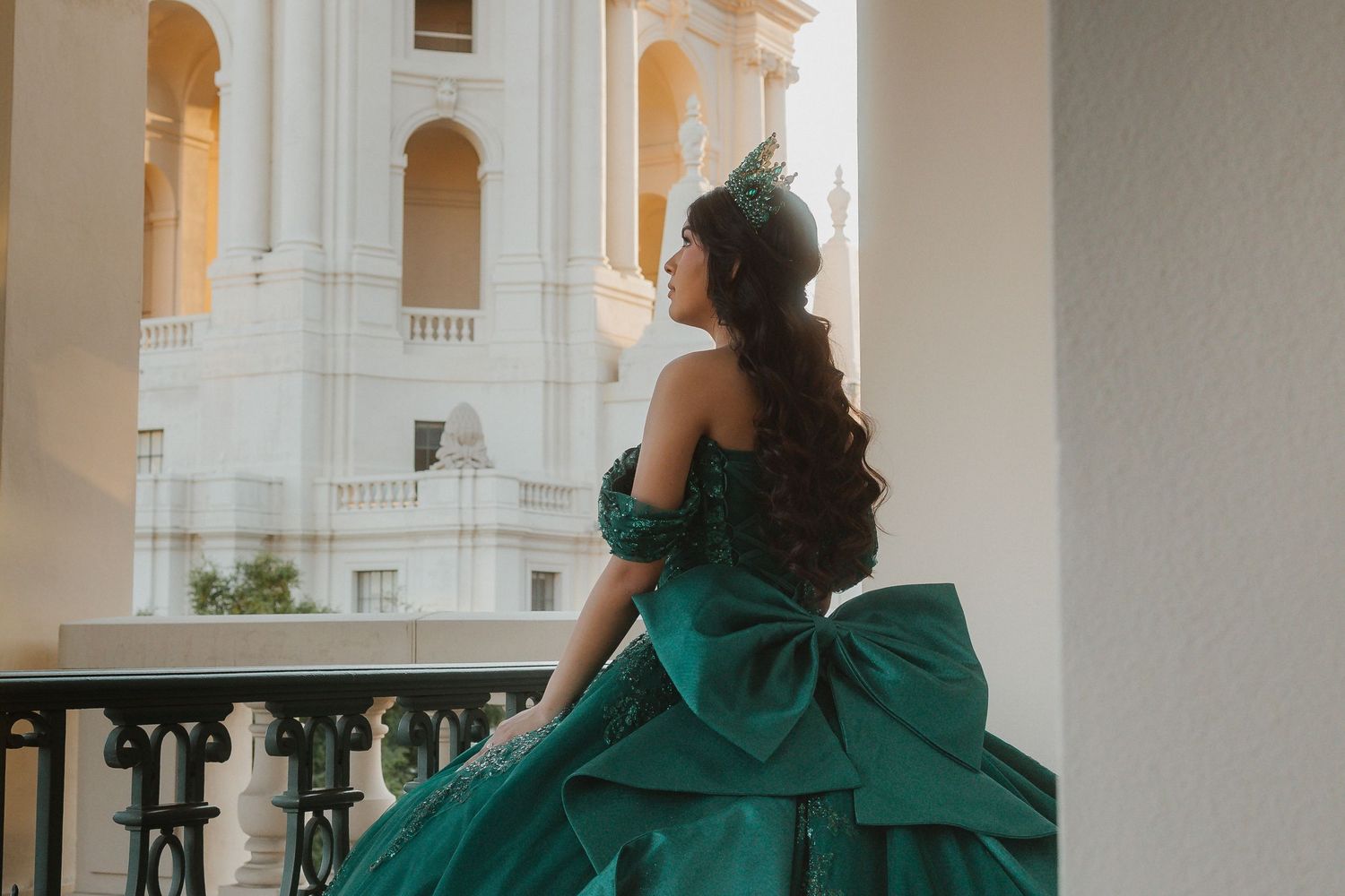 A person in an emerald green gown stands on a white marble balcony gazing at the view of surrounding architecture.