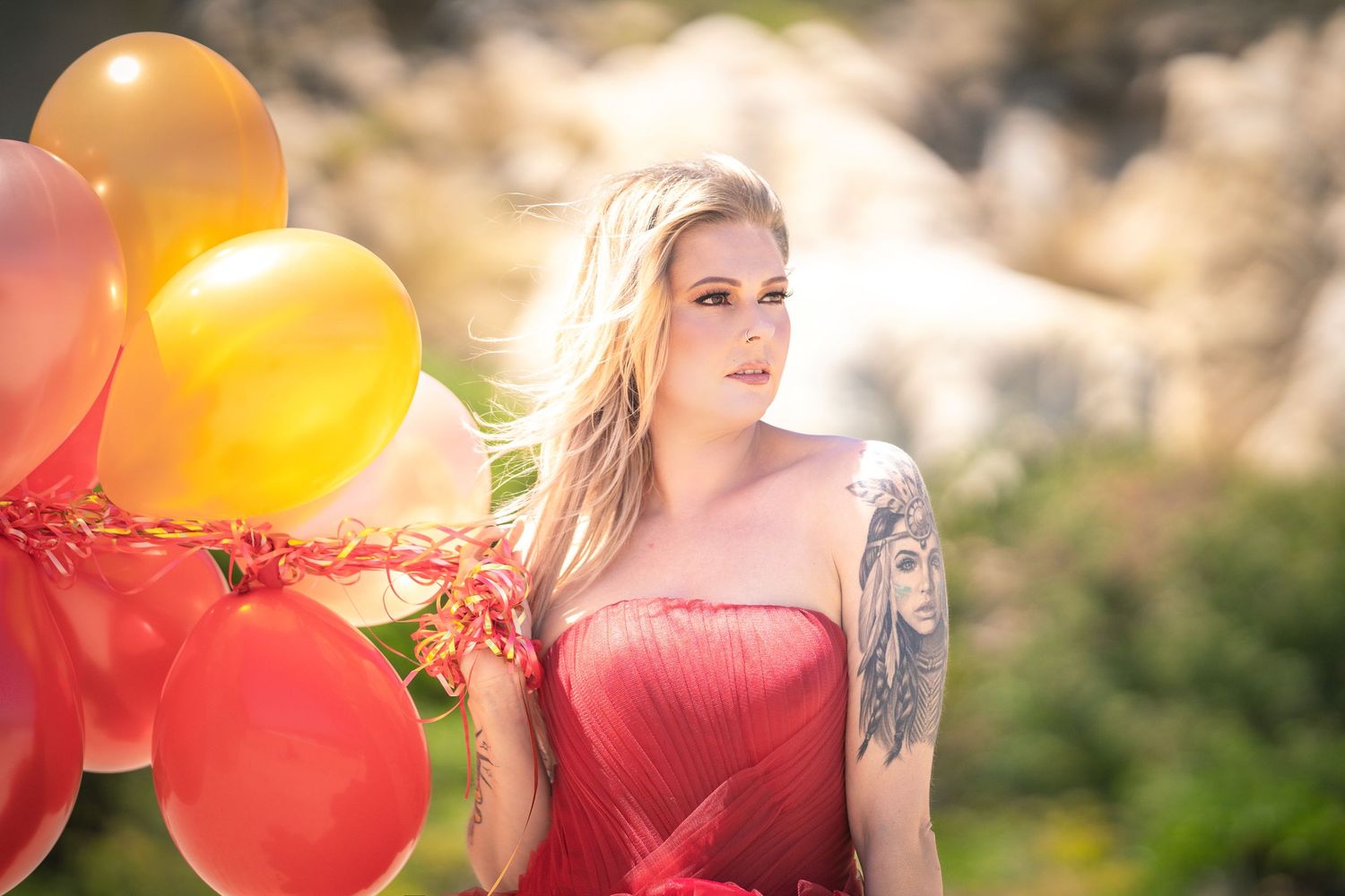 A woman in a flowing pink dress holds vibrant red and orange balloons in a sunny outdoor setting.