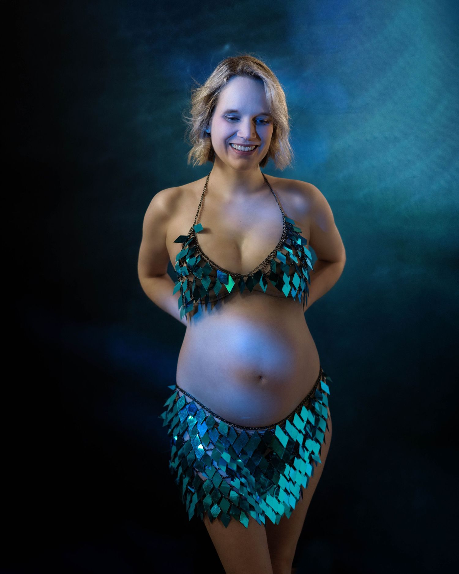 A pregnant figure in a shimmering teal scale-like costume poses against a dark blue backdrop with dramatic lighting.