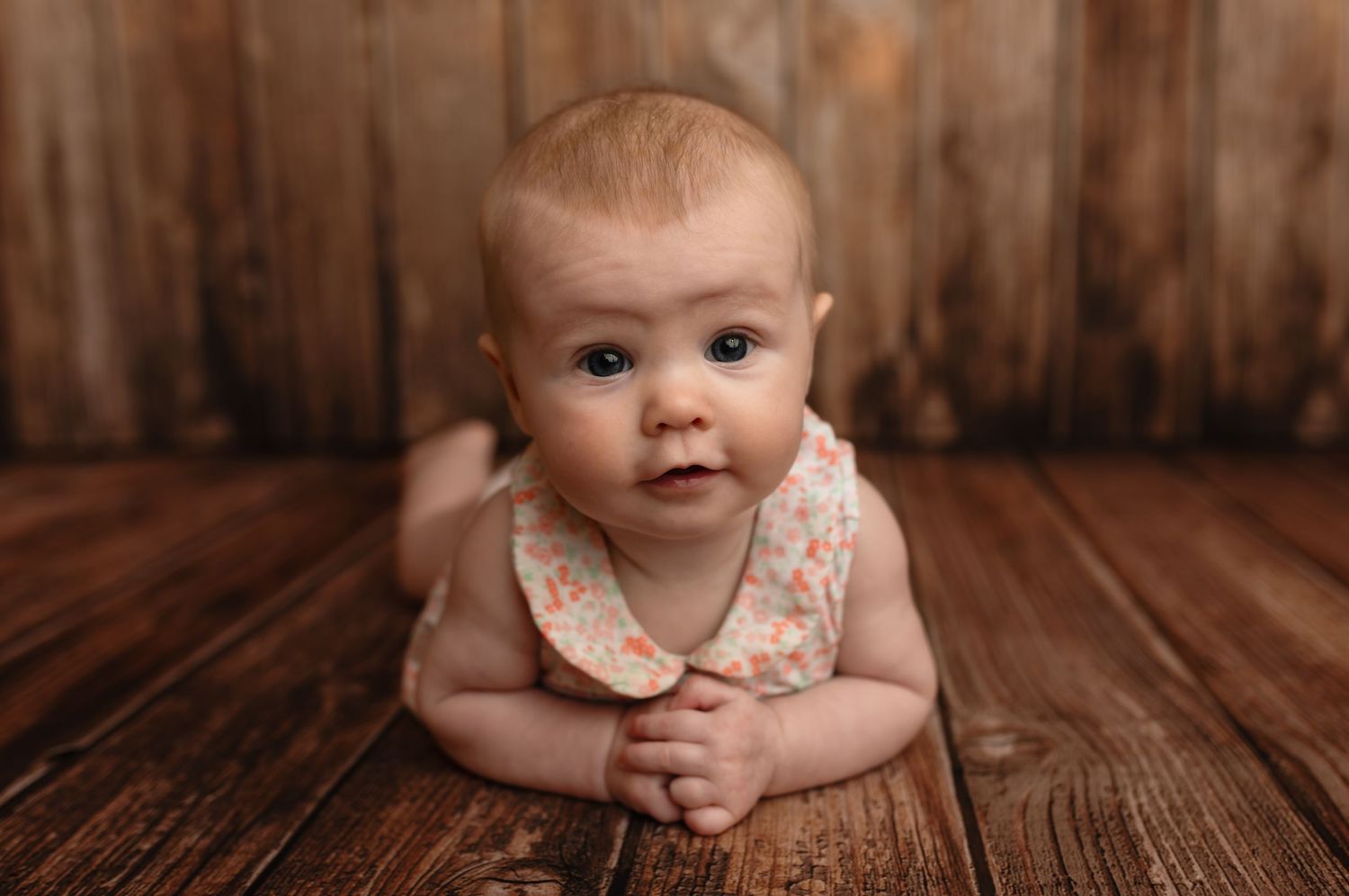 Baby girl smiles at the camera as she sits on warm wood planks in simply photography studio milestone setup by Charlottesville photographer.