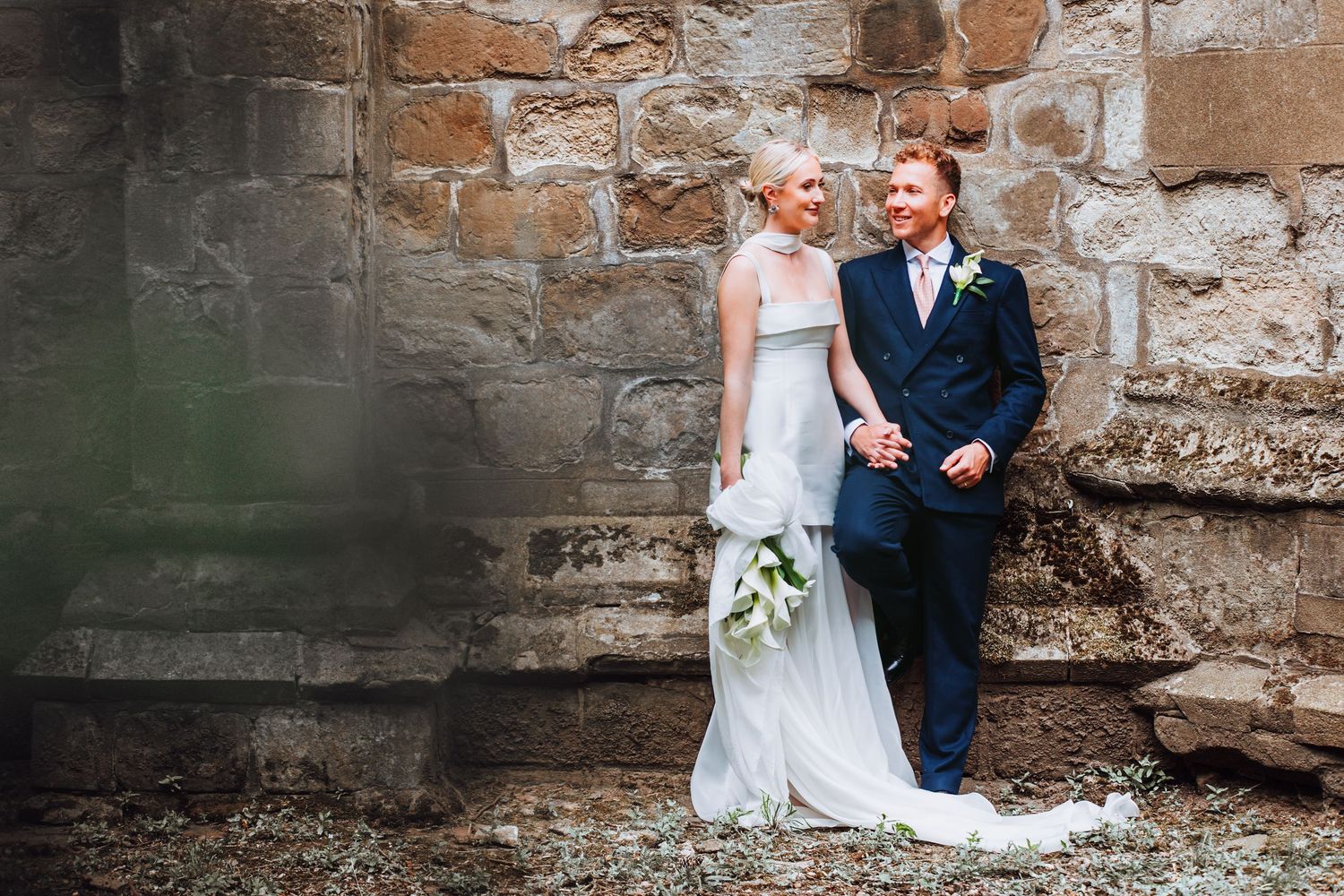 Couple shares candid moments at their rustic outdoor wedding against historic stone wall and garden setting.