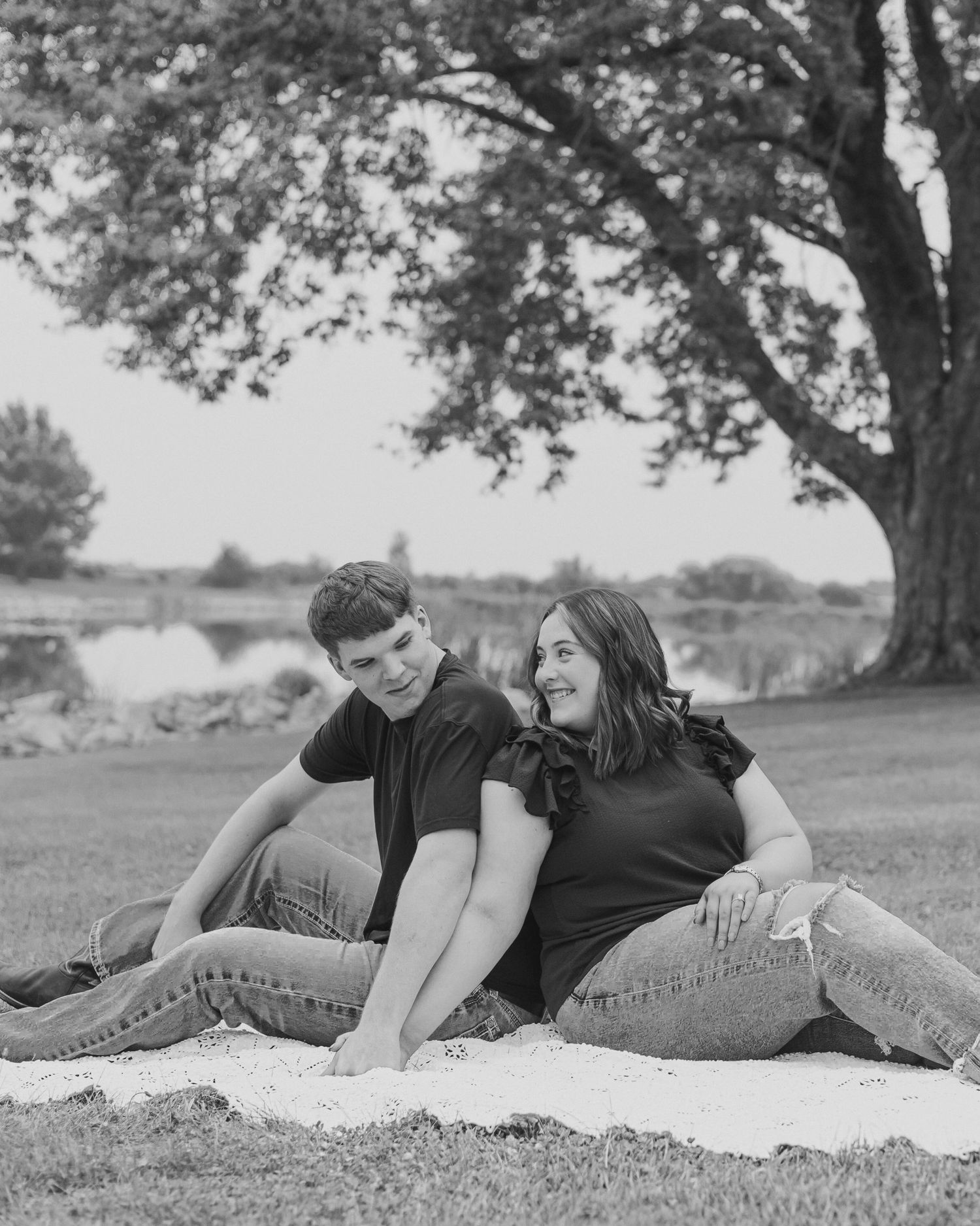 A couple relaxes on a blanket under a large tree by a lake in black and white photography.