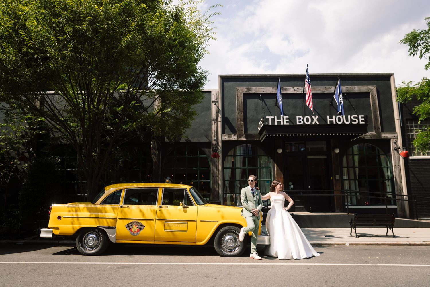 A couple poses with a vintage yellow checker taxi cab in an urban street with green trees.