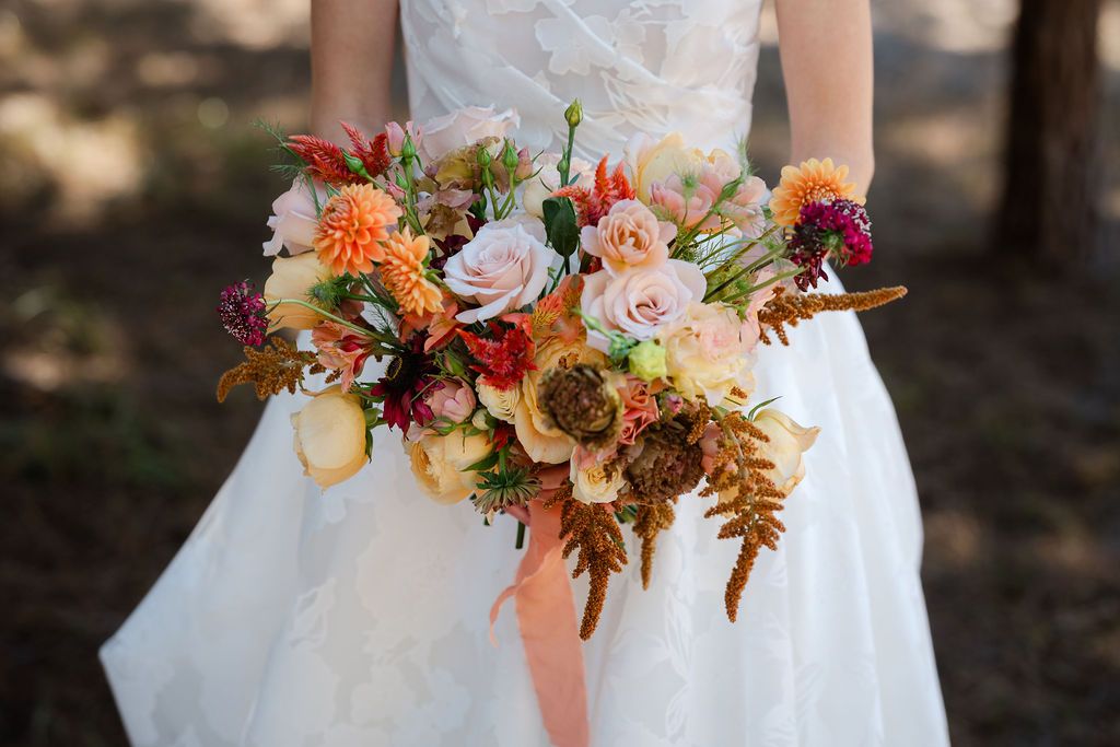 A person in a white wedding gown holds a vibrant fall-colored bridal bouquet with orange and pink flowers.