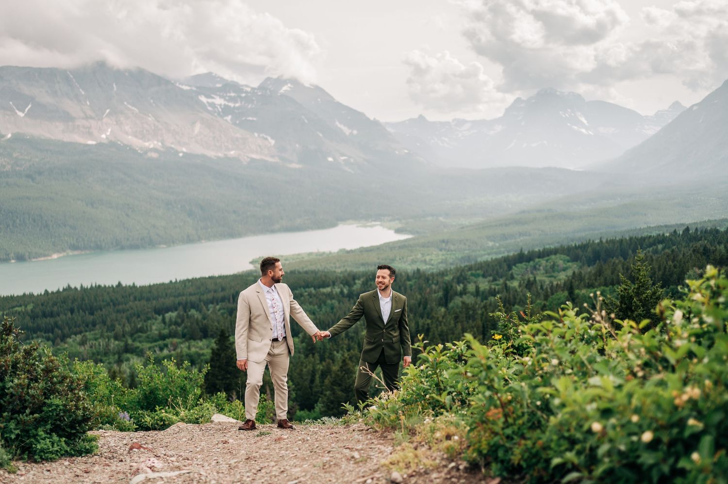 Two people admire a scenic mountain vista with wildflowers in the foreground at Glacier National Park.
