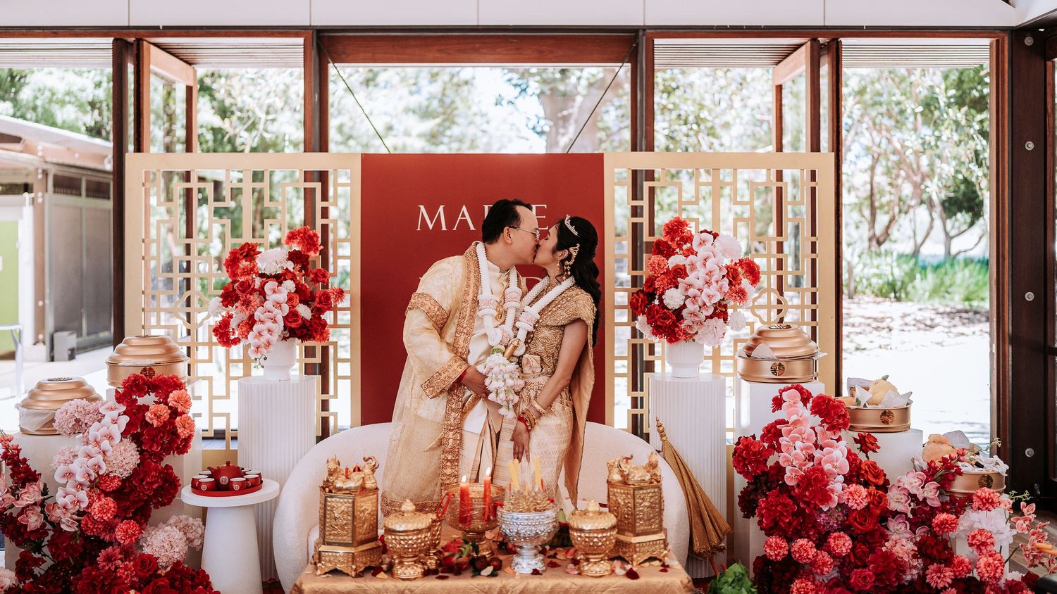 Elegant wedding ceremony setup with red floral arrangements and gold decor against a burgundy backdrop.