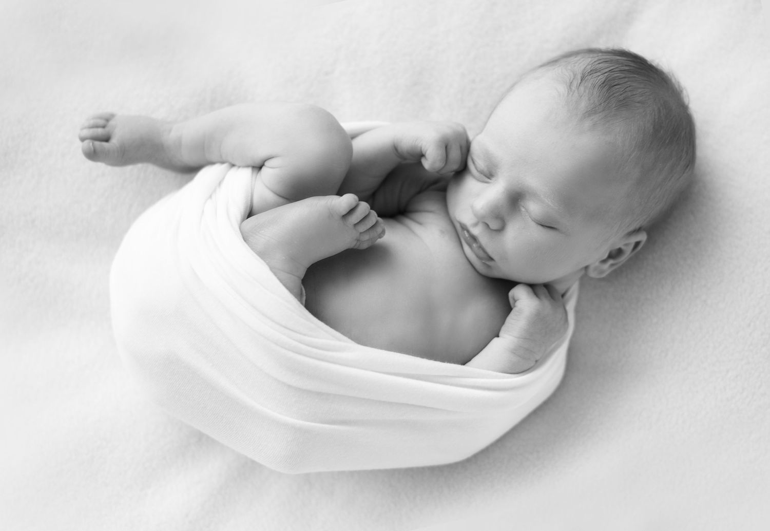 A sleeping newborn baby swaddled in white fabric lies peacefully against a white background.