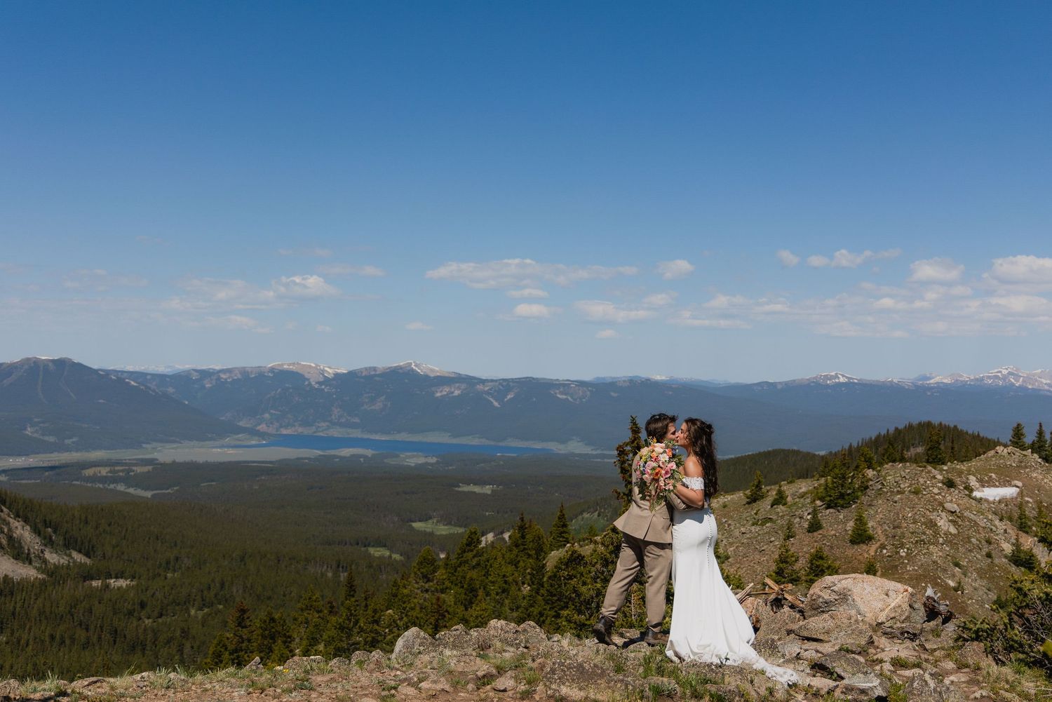 A couple in wedding attire stands on a mountain overlook with sweeping views of forested peaks and valleys below.