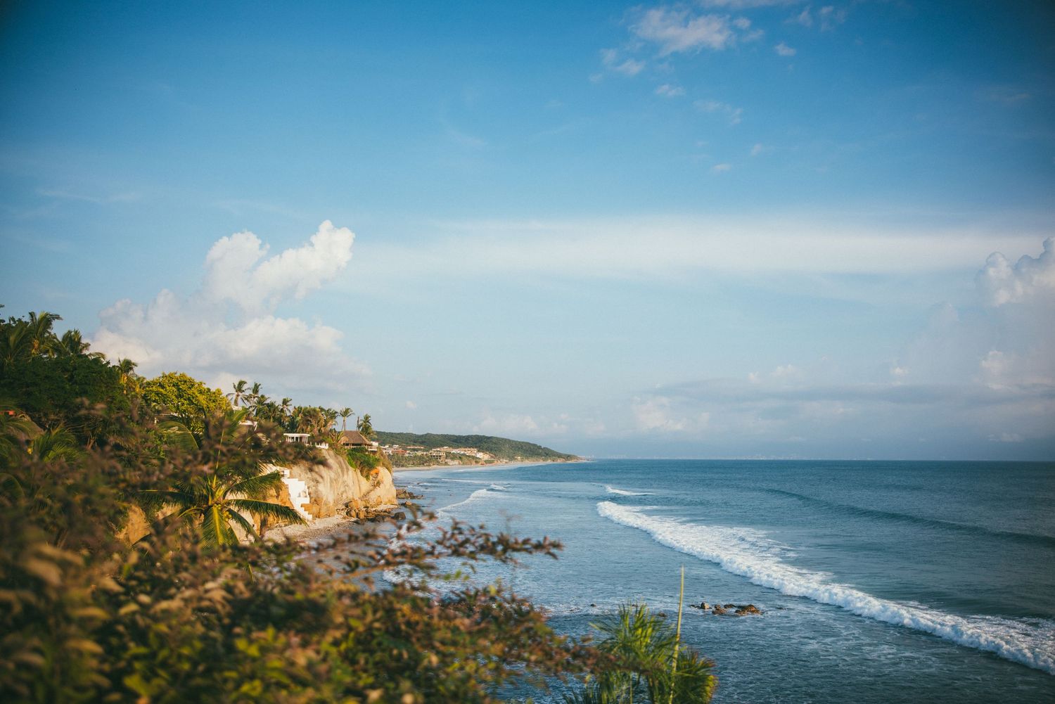 Panoramic view of a tropical coastline with turquoise ocean waves crashing against rocky cliffs under blue skies.