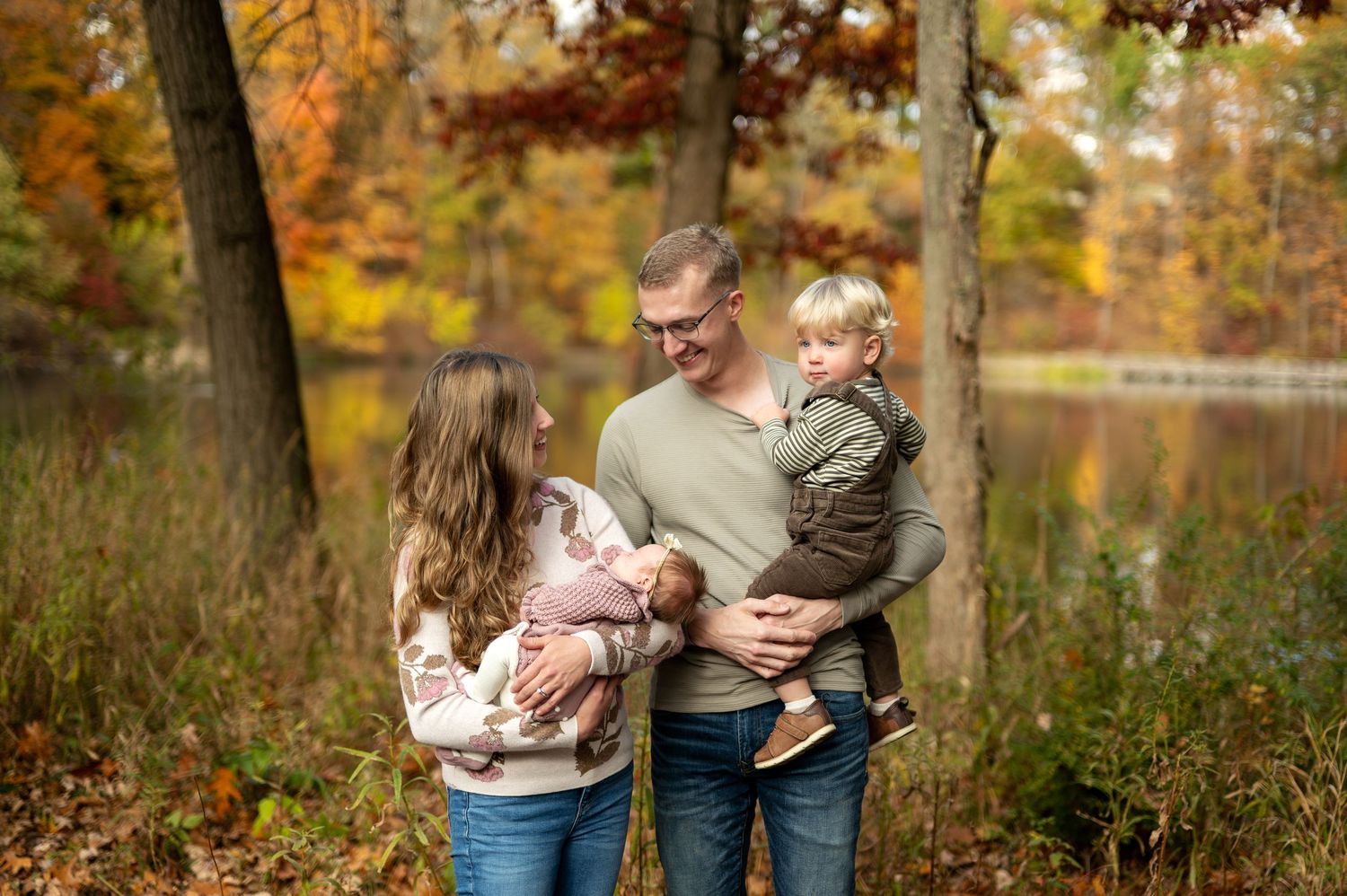 Family of four with newborn and toddler smiling together near a pond in fall foliage.