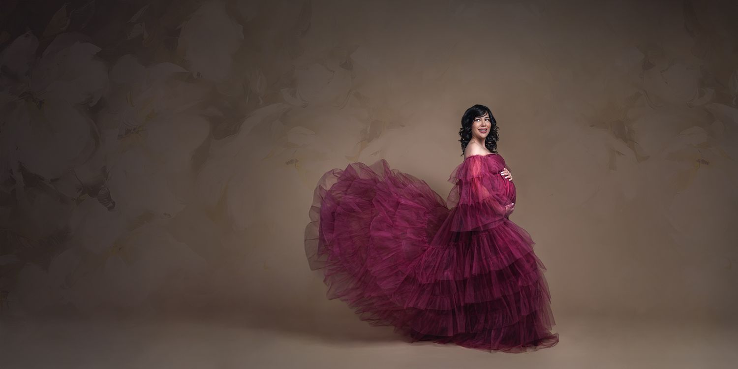Denver maternity photography session of an expectant mother in a beautiful flowing pink gown in front of a floral backdrop in studio.