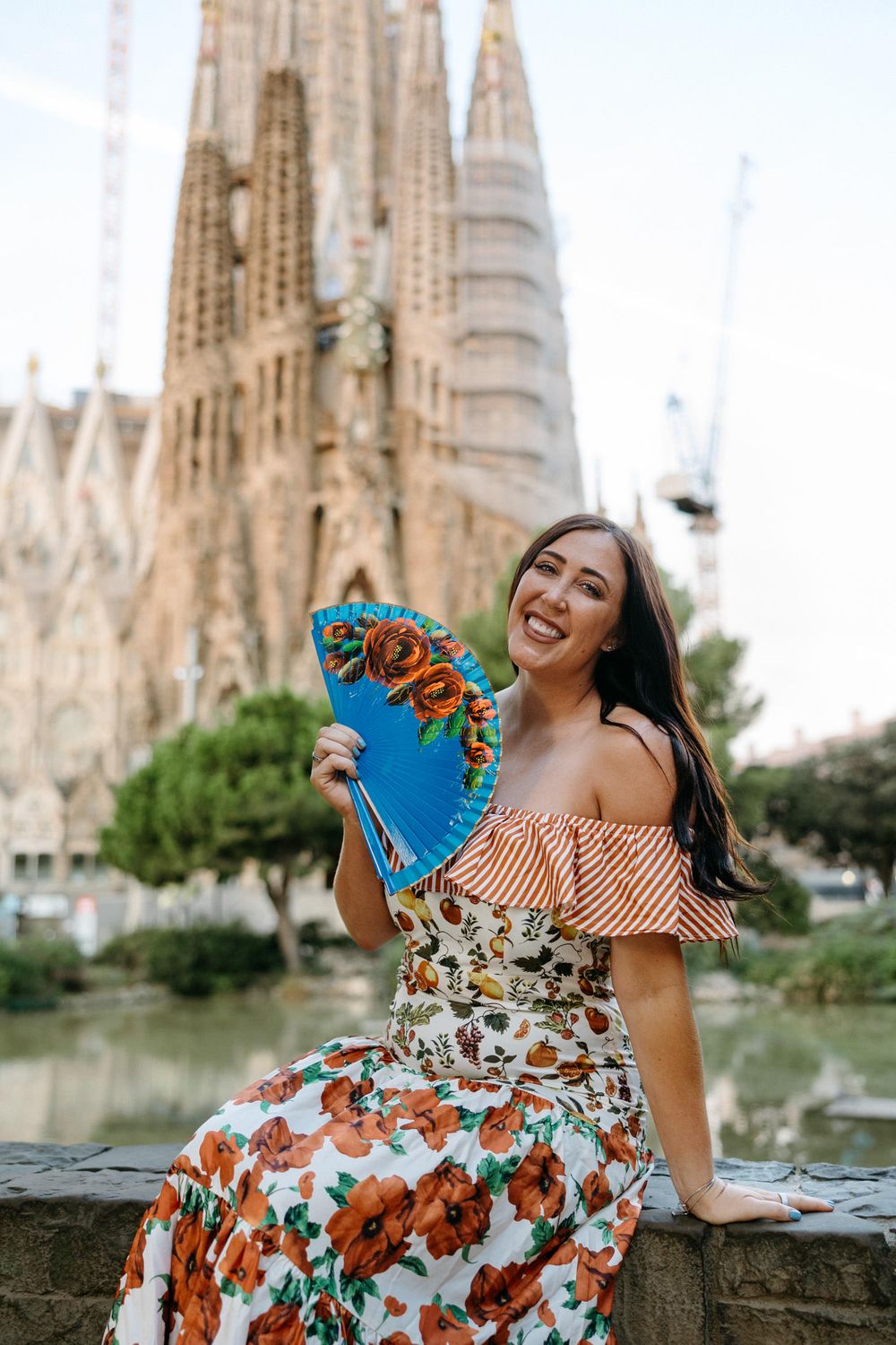 Woman in floral dress sitting on stone wall with Sagrada Familia cathedral spires in background.