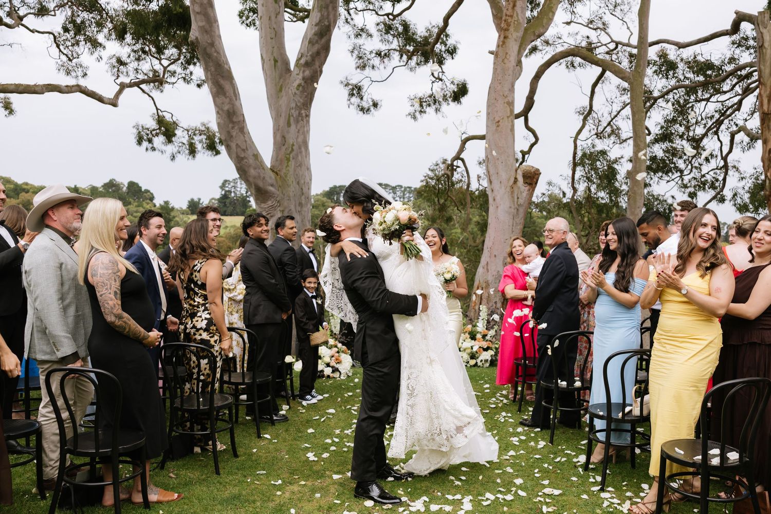 Guests gather outdoors under large trees for a wedding ceremony with white chairs arranged on a grassy lawn.