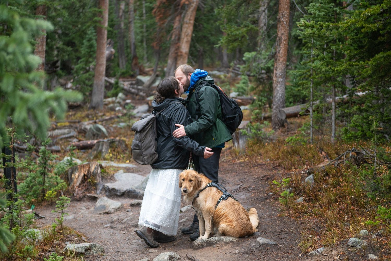A couple on a hiking elopement stop in the trail to share a kiss. They are in wedding attire and raincoats. Their dog sits at their feet.
