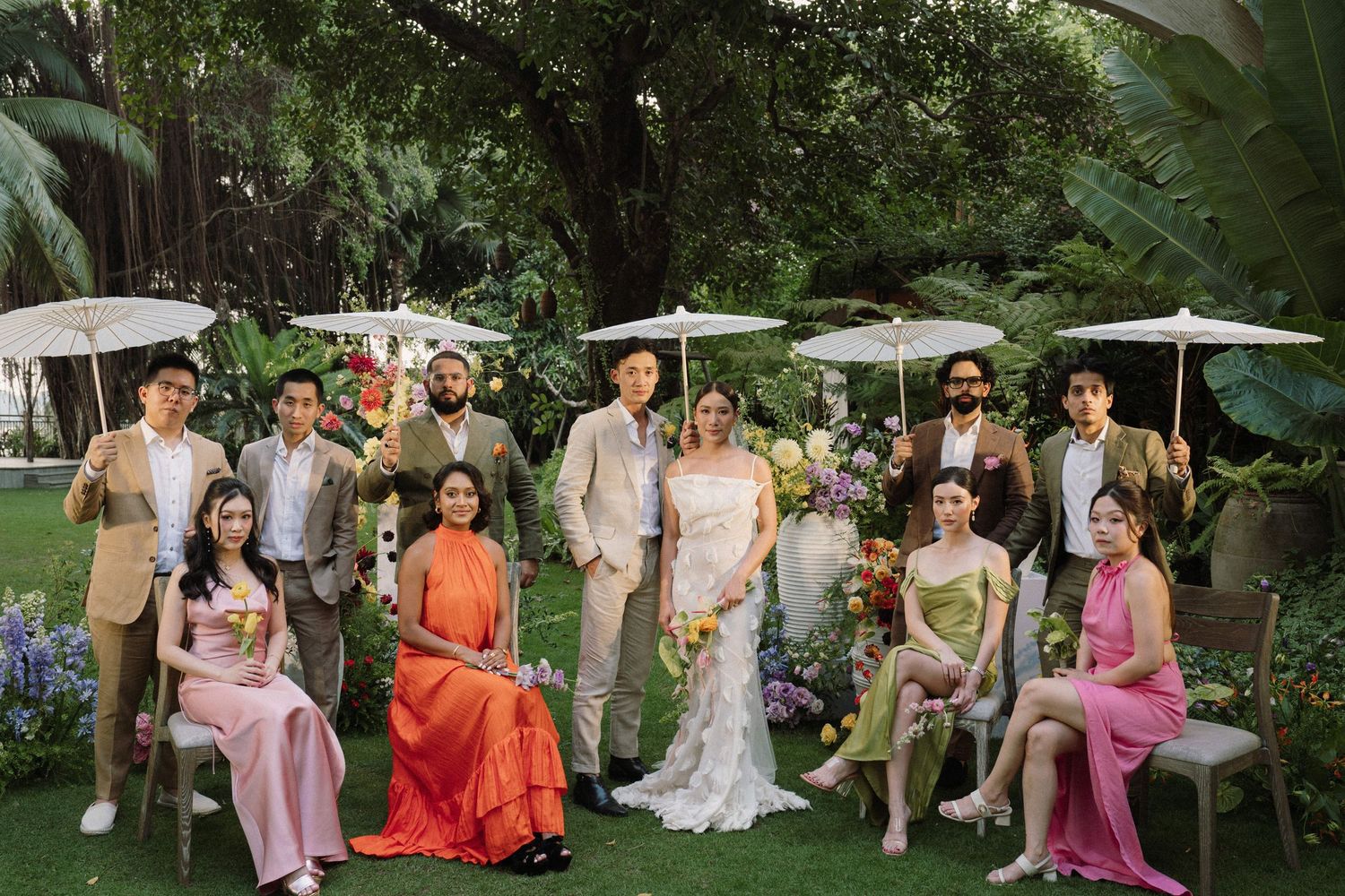 Wedding party in formal attire poses together on garden path surrounded by lush greenery during outdoor celebration.