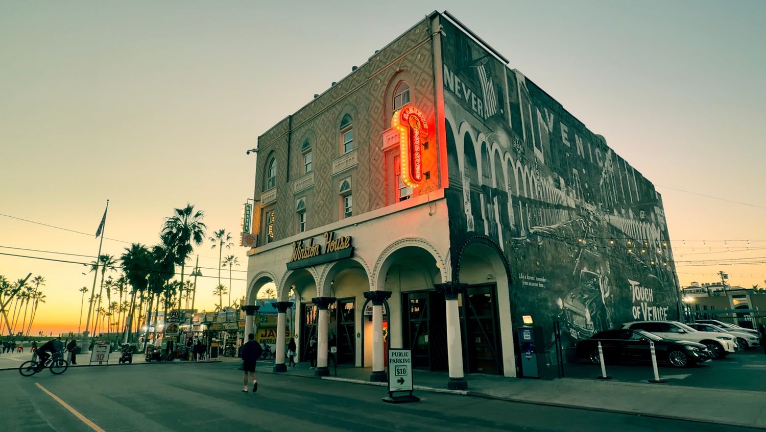 A panoramic view of a modern urban street with palm trees and buildings at sunset against a warm sky.