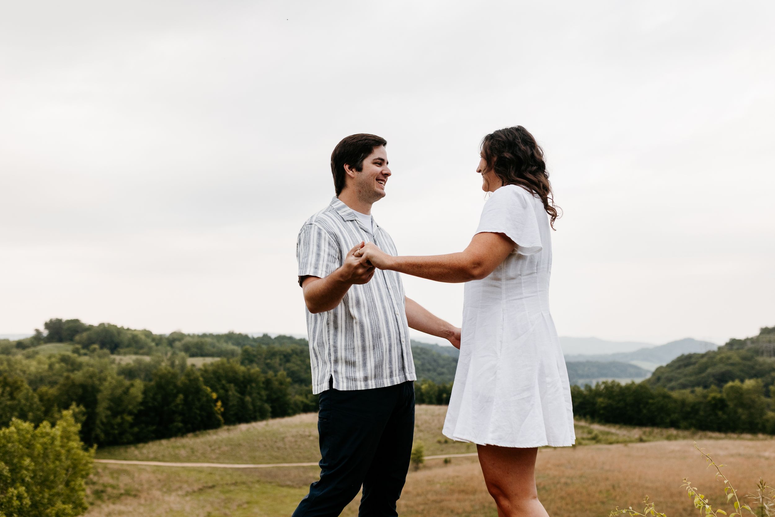 Two people in white and striped outfits embrace in a scenic field on a cloudy summer day.