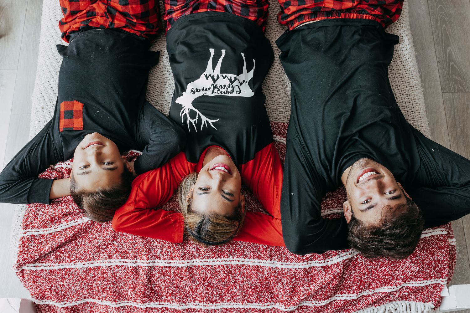 Three people lying down wearing red and black buffalo plaid and dark hoodies on a red and white patterned blanket.