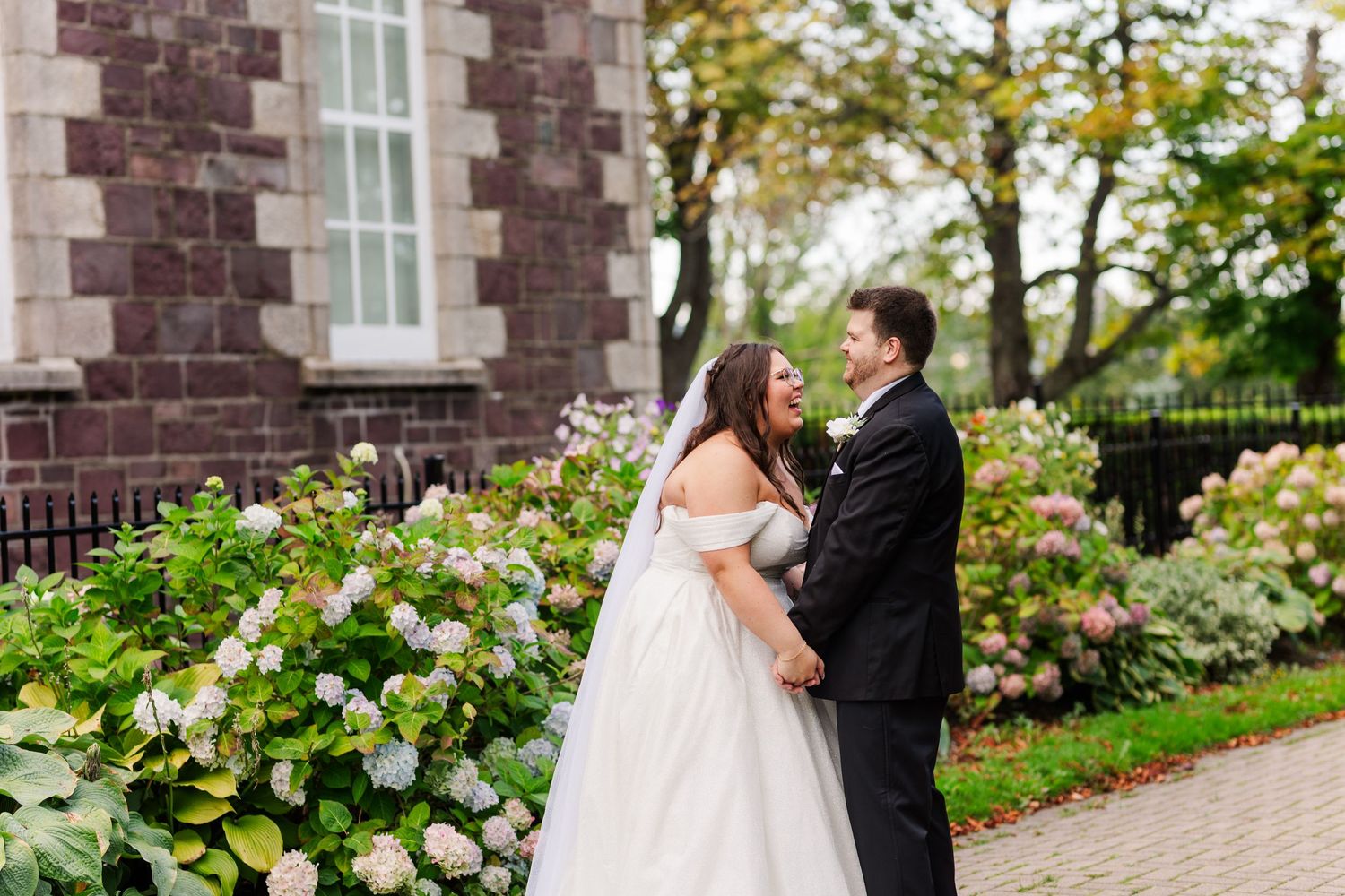 A wedding couple shares a loving embrace while standing on a brick-lined garden path surrounded by autumn trees.