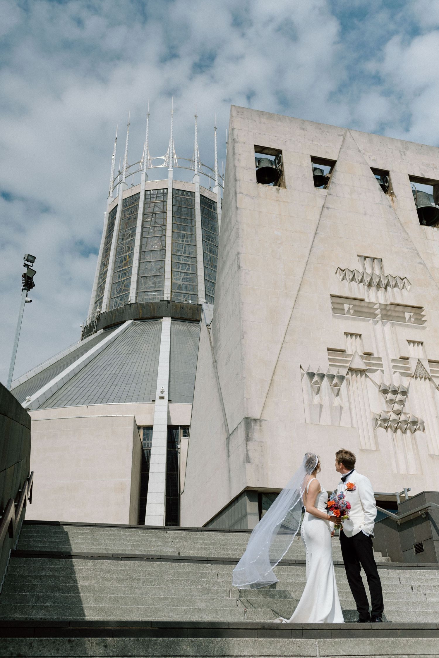 A couple in wedding attire stands on the steps of the Liverpool Metropolitan Cathedral under a cloudy sky.