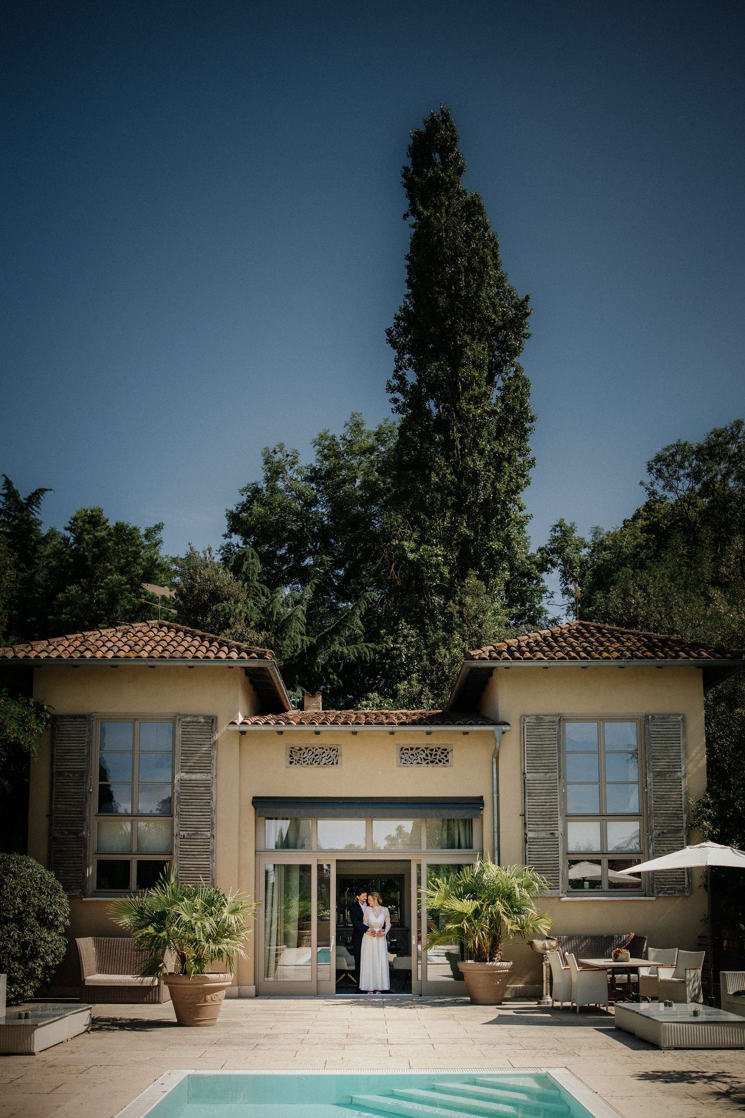 Modern poolside villa with terracotta roof and large windows surrounded by lush landscaping.
