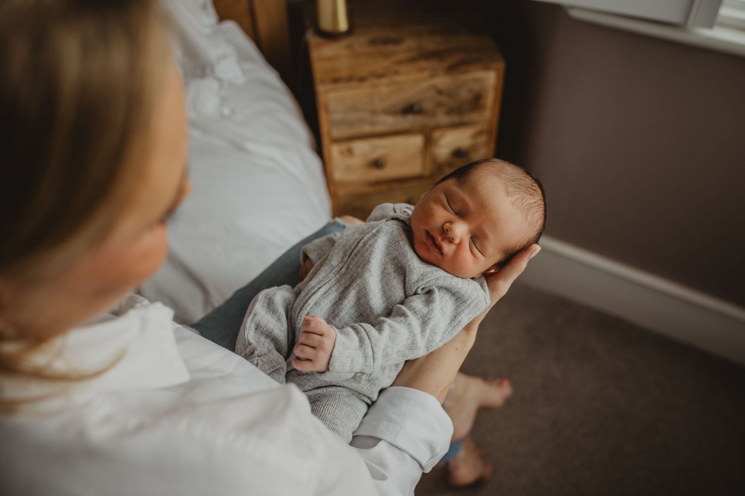 A newborn baby in gray sleepwear rests peacefully against someone wearing white in a cozy bedroom setting.