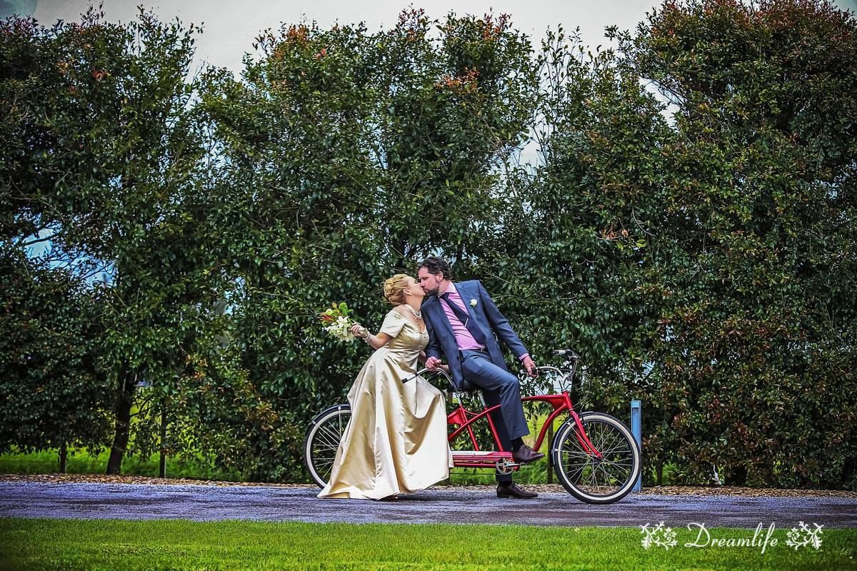 A couple rides a vintage tandem bicycle through lush countryside gardens under dramatic cloudy skies.