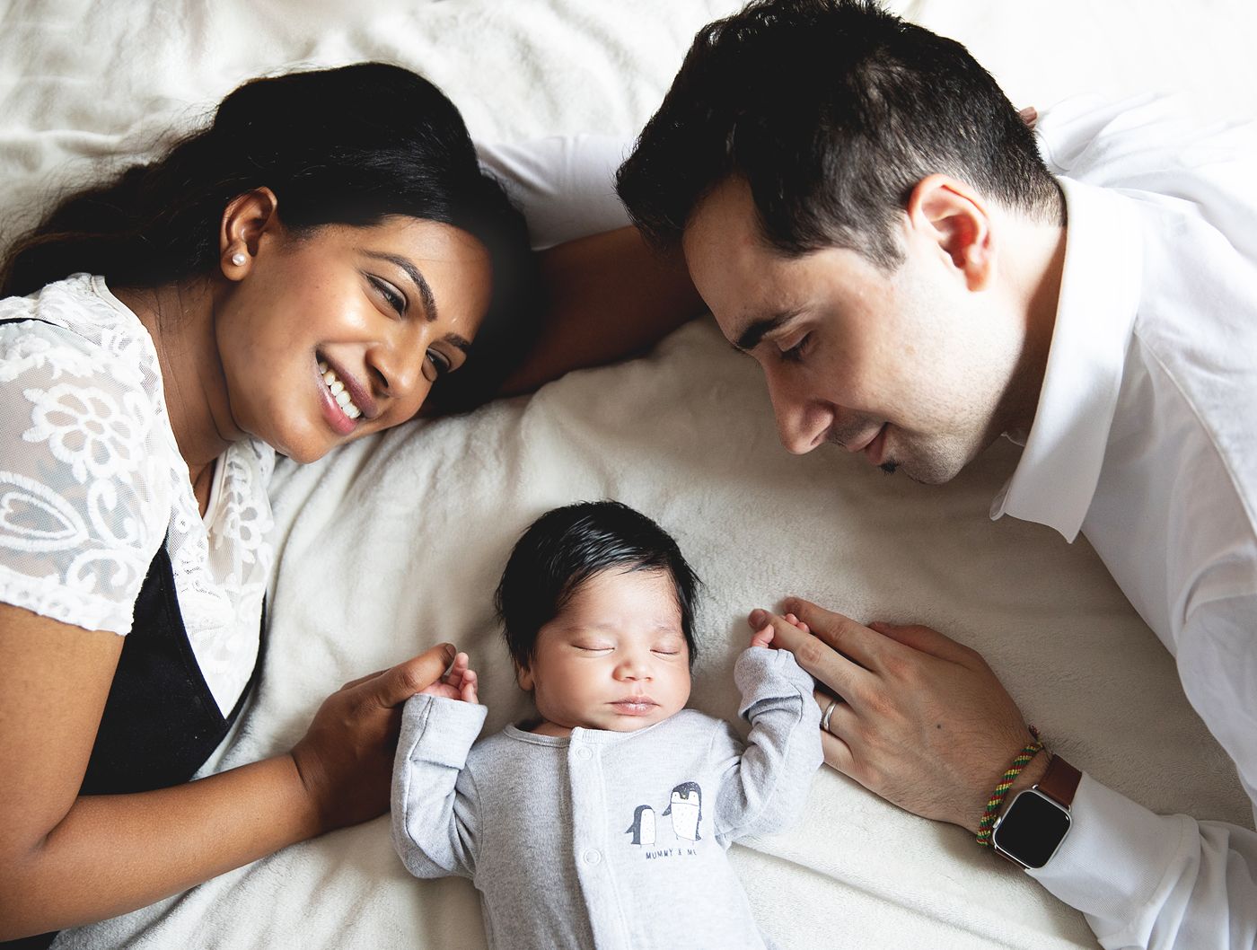 A happy couple lying on a bed smiling at their sleeping newborn baby dressed in a grey onesie. At a home newborn session in bexley