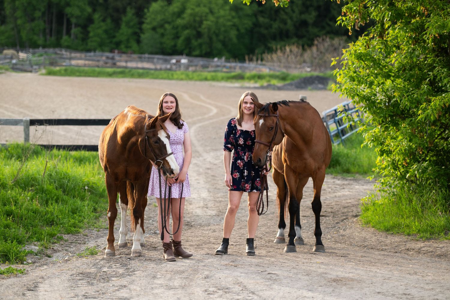Group of people walking with several brown horses in an outdoor field setting next to trees and bushes.