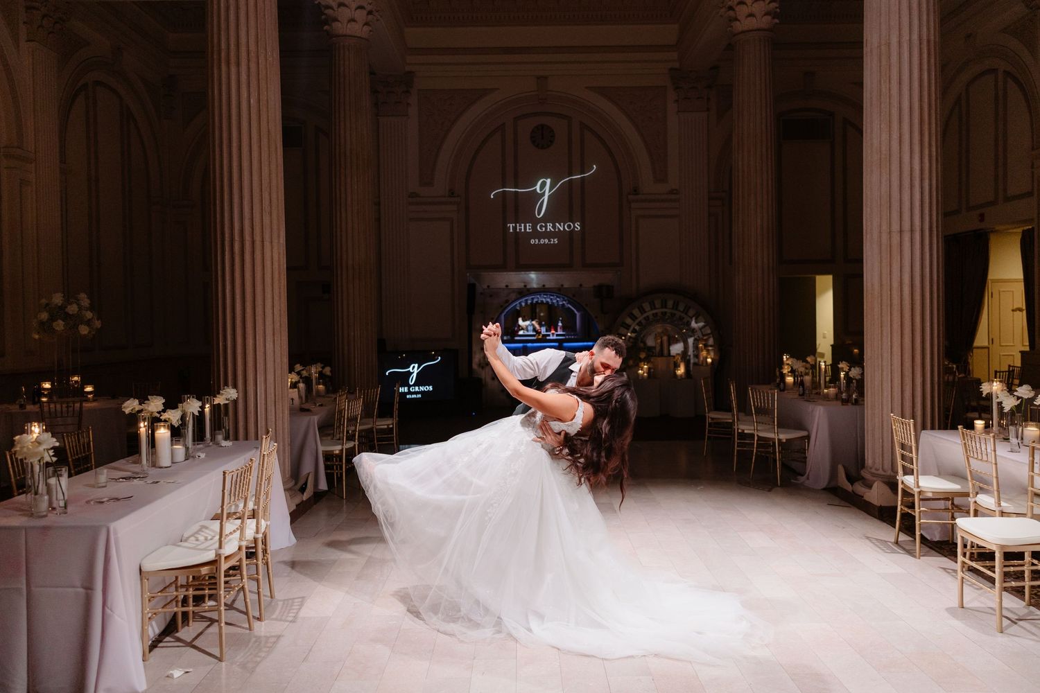 A panoramic view of an elegant wedding ceremony in a white columned ballroom with gold chairs and floral decorations.