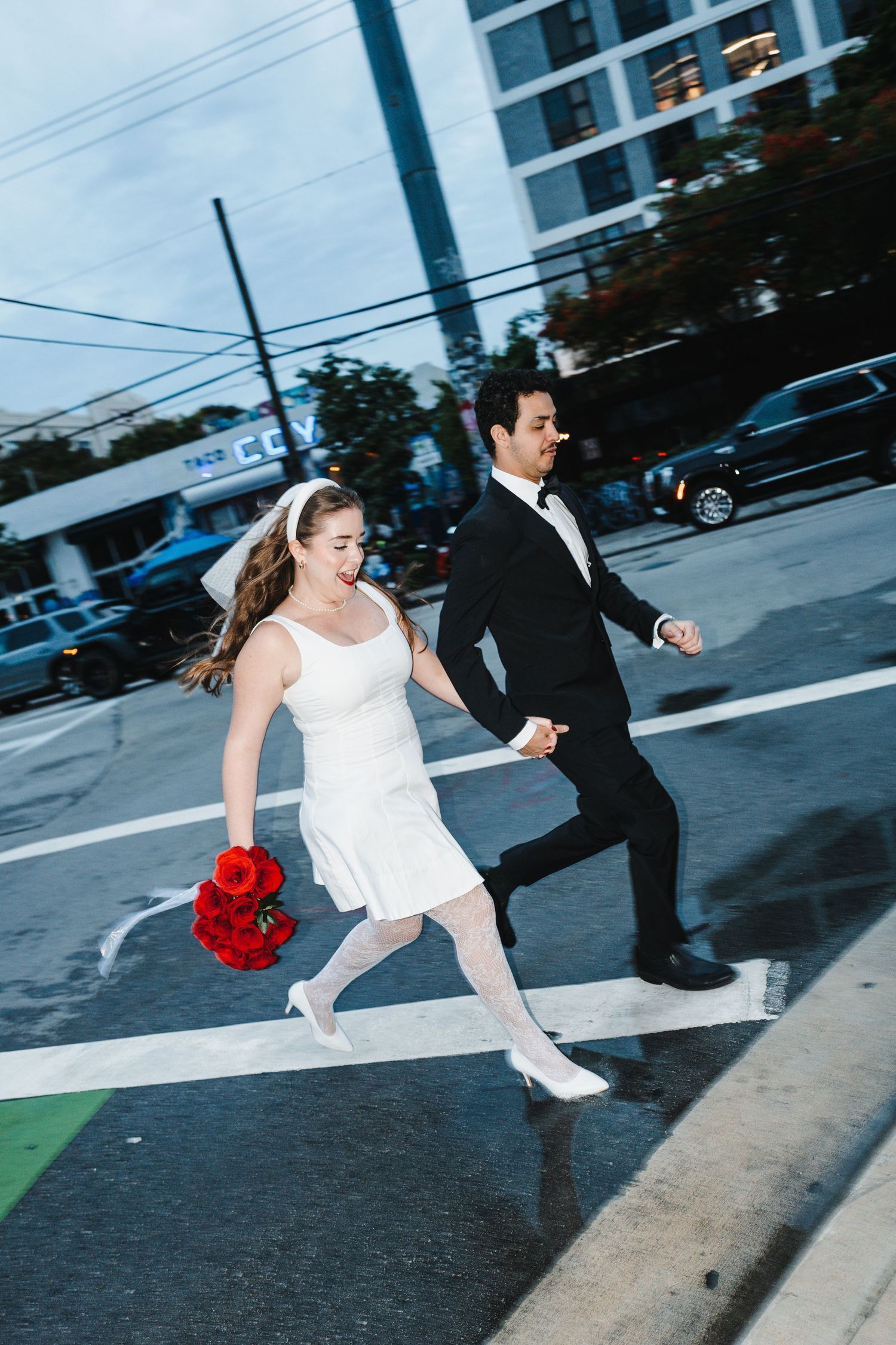 A couple in wedding attire runs across a city crosswalk while holding a red bouquet.