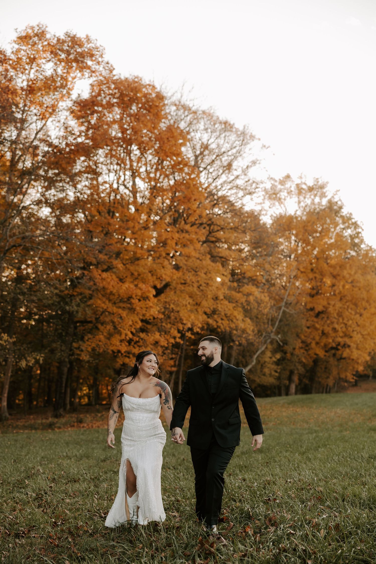 A couple in wedding attire walks hand in hand through a grassy field surrounded by autumn trees with orange foliage.