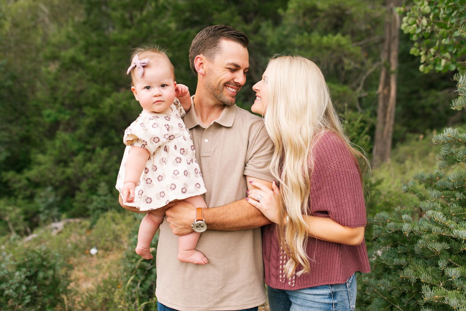 Happy young family with baby girl posing outdoors in a mountain meadow surrounded by tall pine trees.
