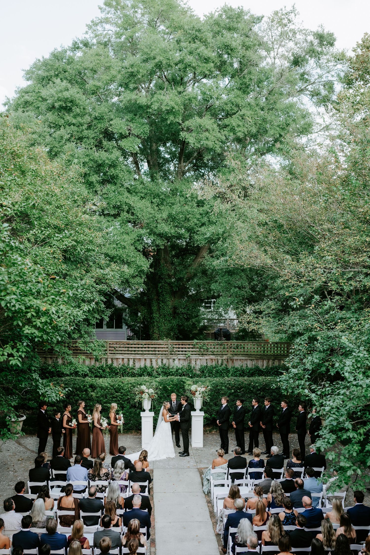 Outdoor garden wedding ceremony with bridal party at altar, guests seated on white chairs, surrounded by lush green trees.