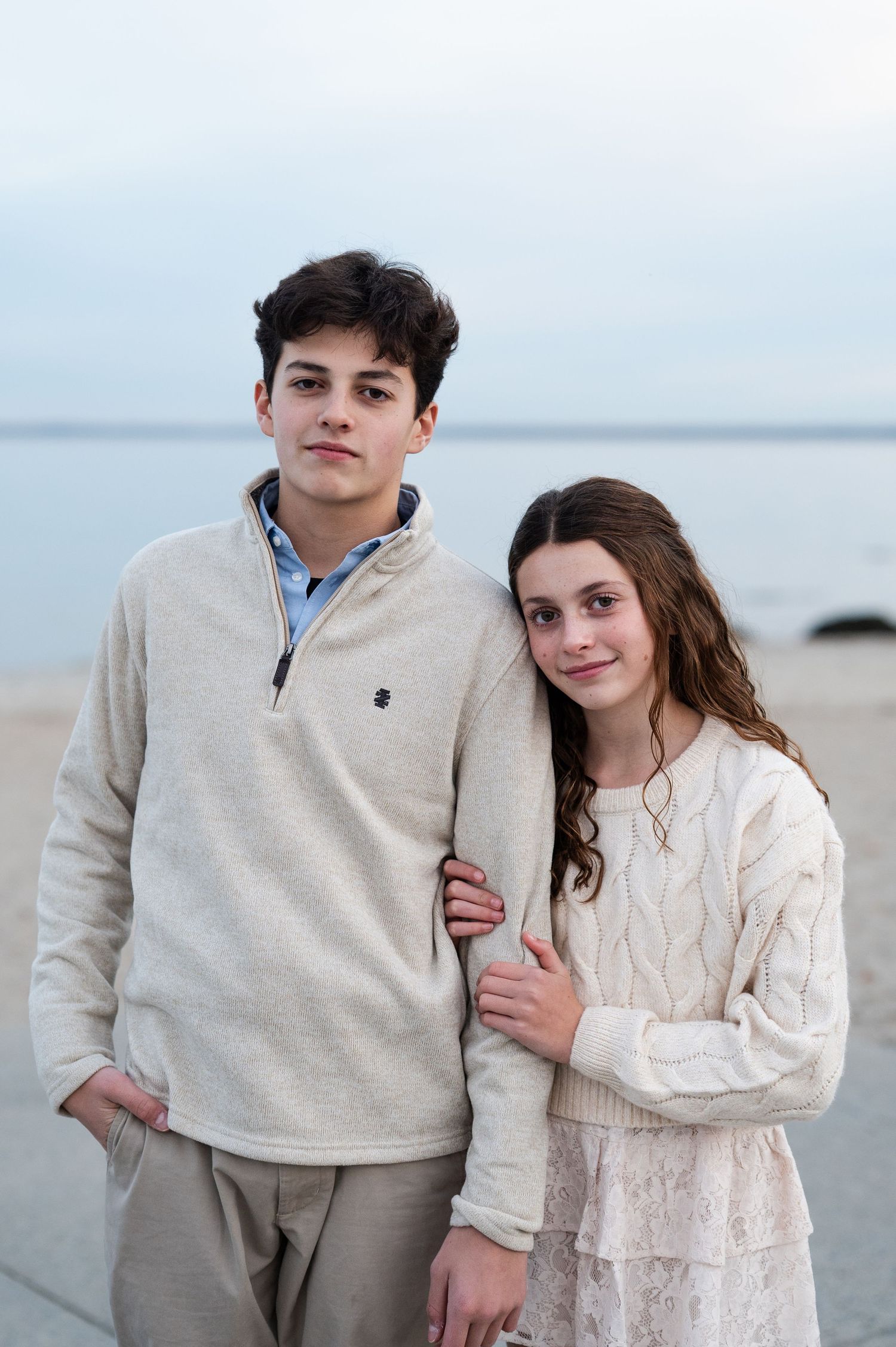 A brother and sister laughing and embracing on a sandy beach with calm ocean waters in the background.