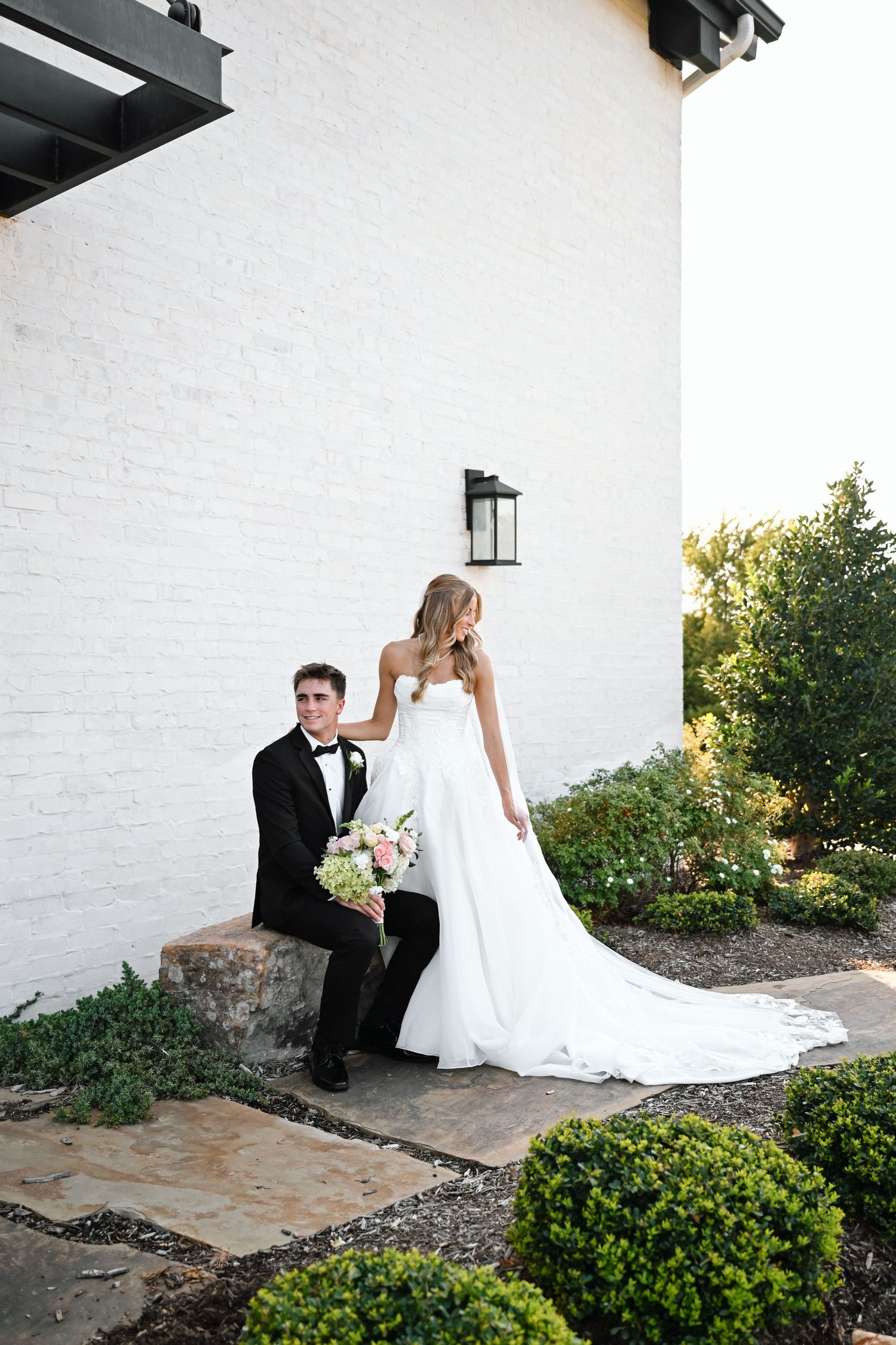 Bride in white gown holds bouquet beside groom in tuxedo seated on stone steps outside white brick building.